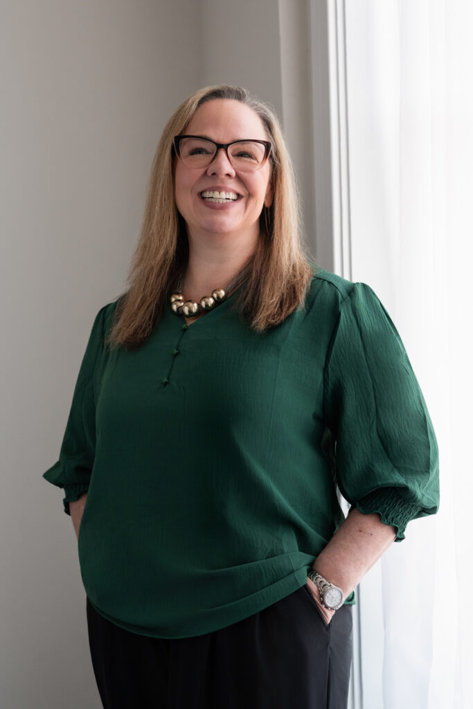 Lynn Cross standing by a window in a green blouse, smiling